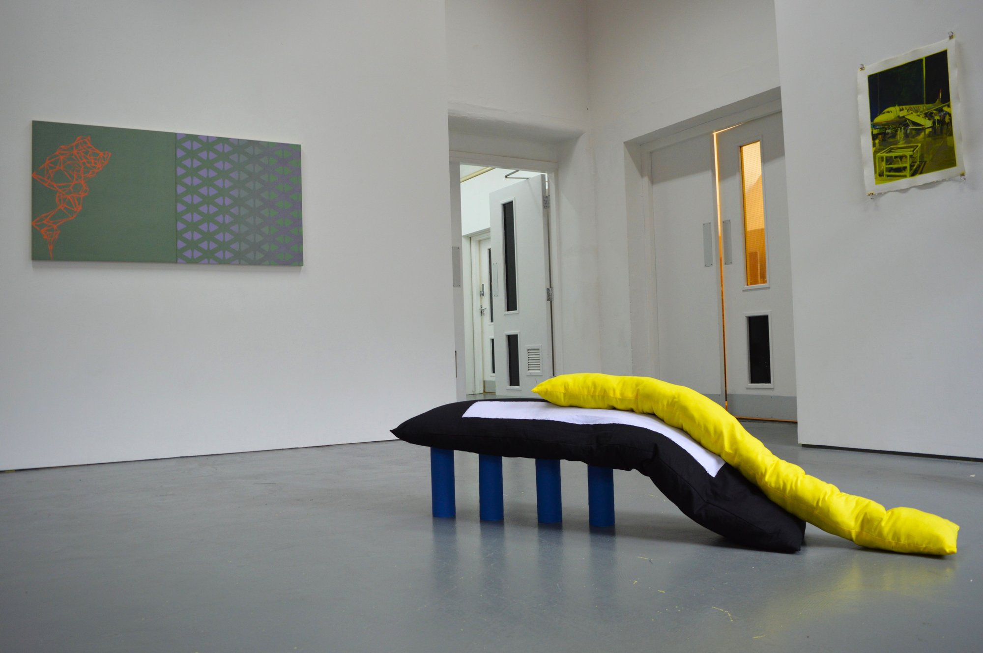 Soft sculpture piece with 4 tubular blue legs, a long black fabric stuffed piece with tube yellow placed on top.