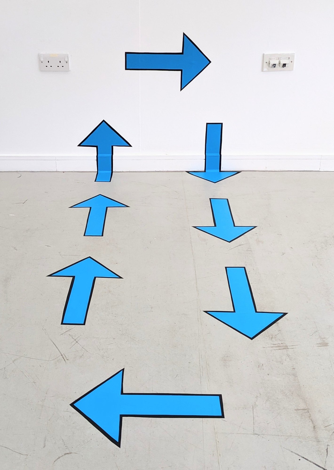 8 blue arrows on the ground and going up blank white gallery wall. Each arrow has a black border and is made from vinyl.